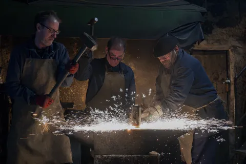 Tres herreros en taller de forja tradicional trabajando el metal con martillo y yunque, produciendo chispas incandescentes.
