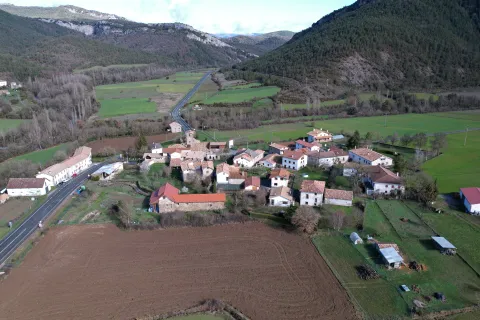 Vista aérea de Guesa con sus casas de piedra dispersas en el valle pirenaico, rodeadas de campos de cultivo y montañas boscosas al fondo.