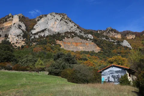 Acantilados calizos de Sierra de Leyre con prado verde y cabaña con mural histórico en primer plano.