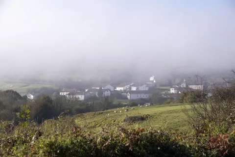 Pueblo rural envuelto en niebla matutina, con caseríos dispersos entre prados y vegetación.