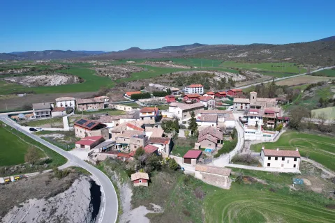 Vista aerea del pueblo de Romanzado con casas dispersas, campos verdes y montana al fondo en el prepirineo.