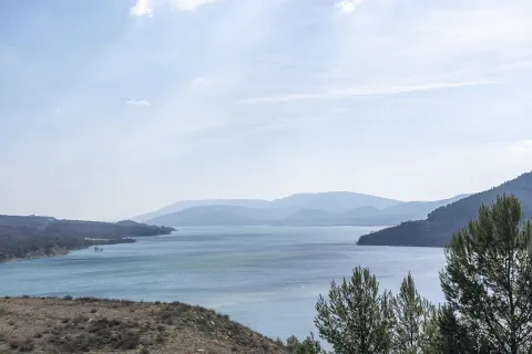 Embalse de Yesa rodeado de montes boscosos y montañas al fondo bajo cielo despejado con nubes ligeras.