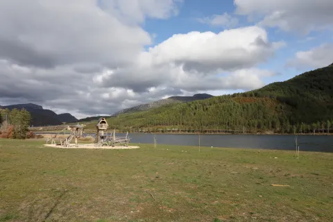 Embalse de Nagore rodeado de bosque pirenaico, con prado en primer plano y parque de madera y montes al fondo bajo cielo nublado.