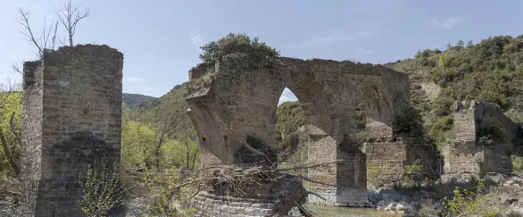 Ruinas de puente medieval con arcos de piedra sobre río en Puente de los Roncaleses, rodeado de vegetación y montaña.
