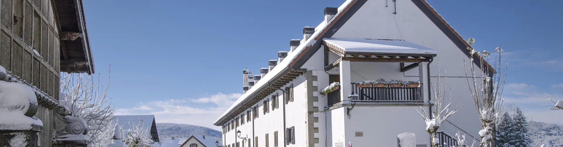Señalización de entrada a Auritz-Burguete con escudos heráldicos cubiertos de nieve y paisaje pirenaico nevado de fondo.