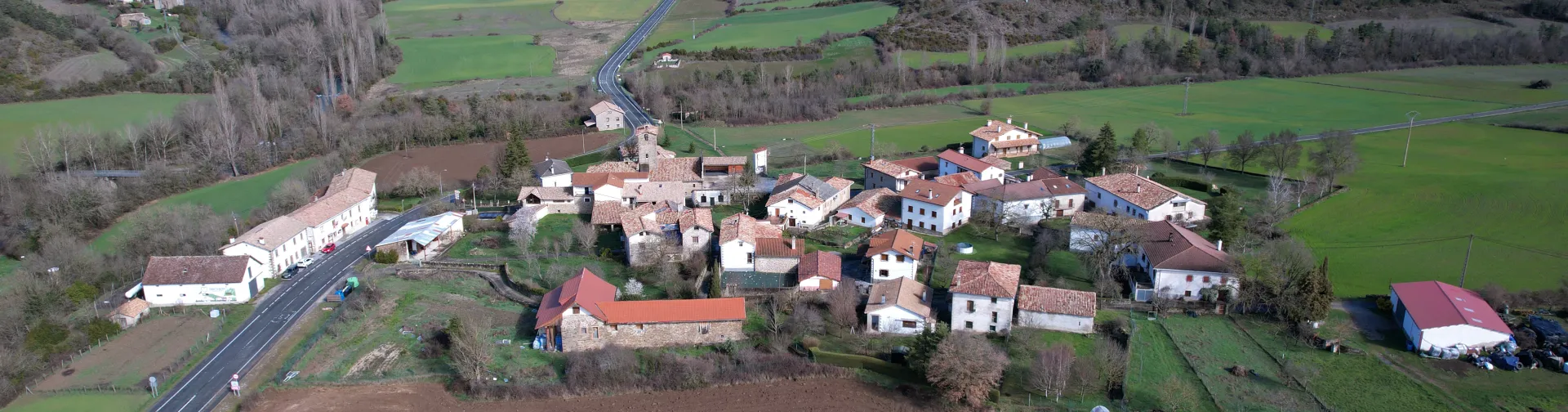 Vista aérea de Guesa con sus casas de piedra dispersas en el valle pirenaico, rodeadas de campos de cultivo y montañas boscosas al fondo.