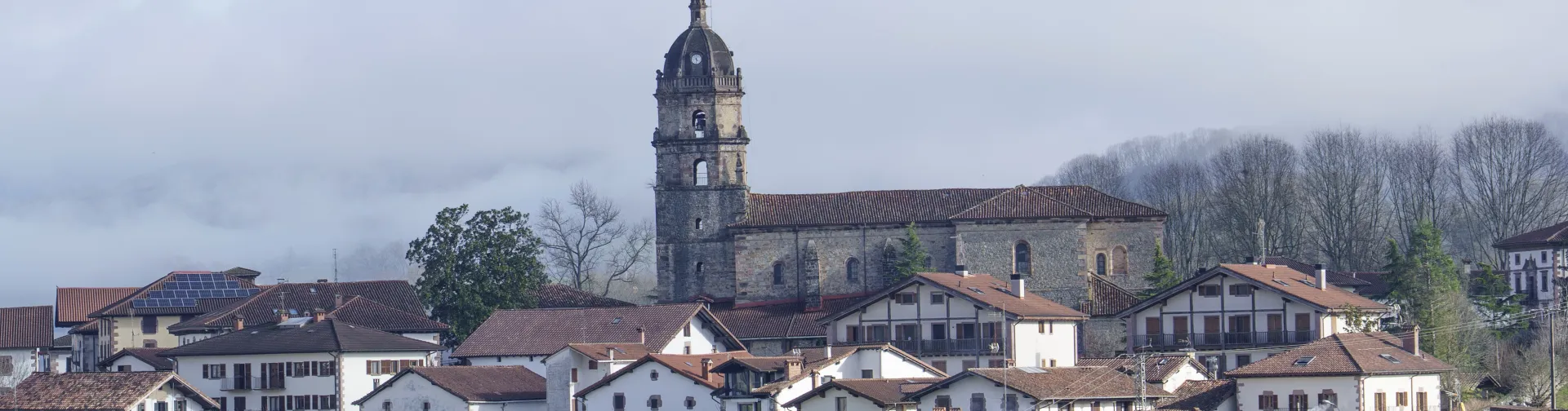 Vista general de Irurita con iglesia de piedra, casas tradicionales blancas y montañas tapadas con las nubes de fondo.