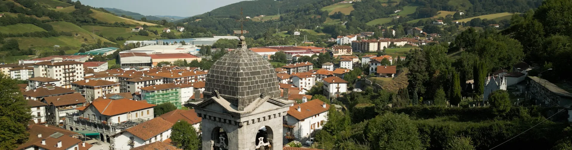 Vista aérea de Leitza con iglesia parroquial de cúpula en primer plano y casas de tejados rojizos entre montes y prados.