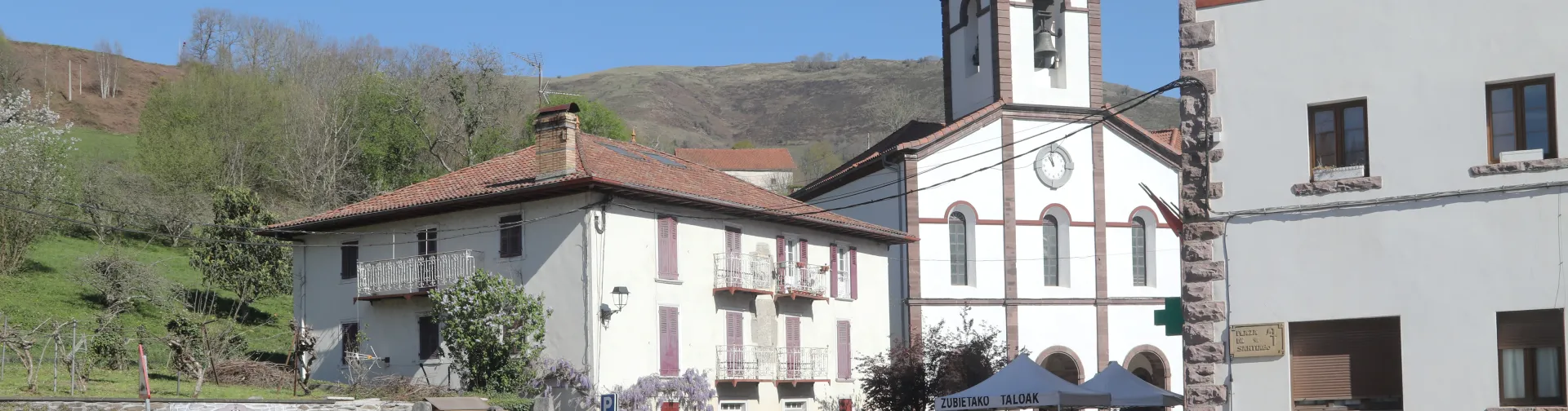 Plaza de Luzaide-Valcarlos con iglesia de arquitectura tradicional, campanario y edificios de piedra, con visitantes y montañas al fondo.