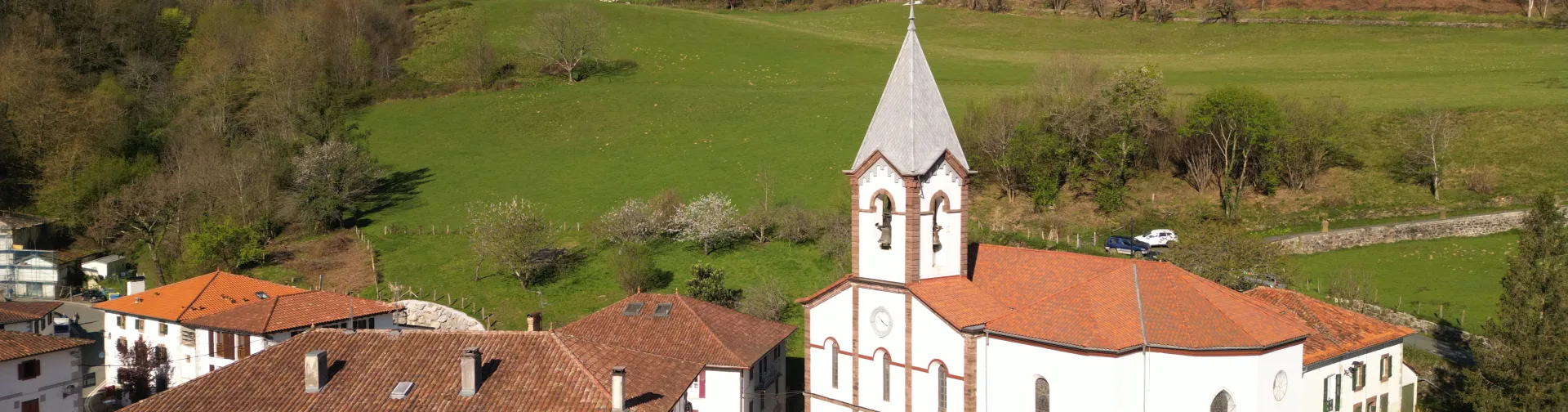 Iglesia blanca con campanario en Luzaide-Valcarlos, rodeada de prados verdes y montañas del Pirineo en valle fluvial.