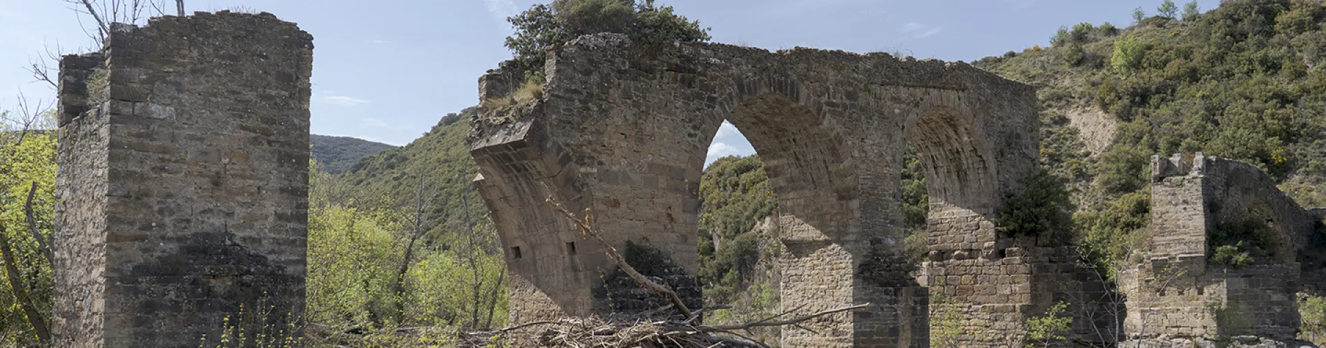 Ruinas de puente medieval con arcos de piedra sobre río en Puente de los Roncaleses, rodeado de vegetación y montaña.