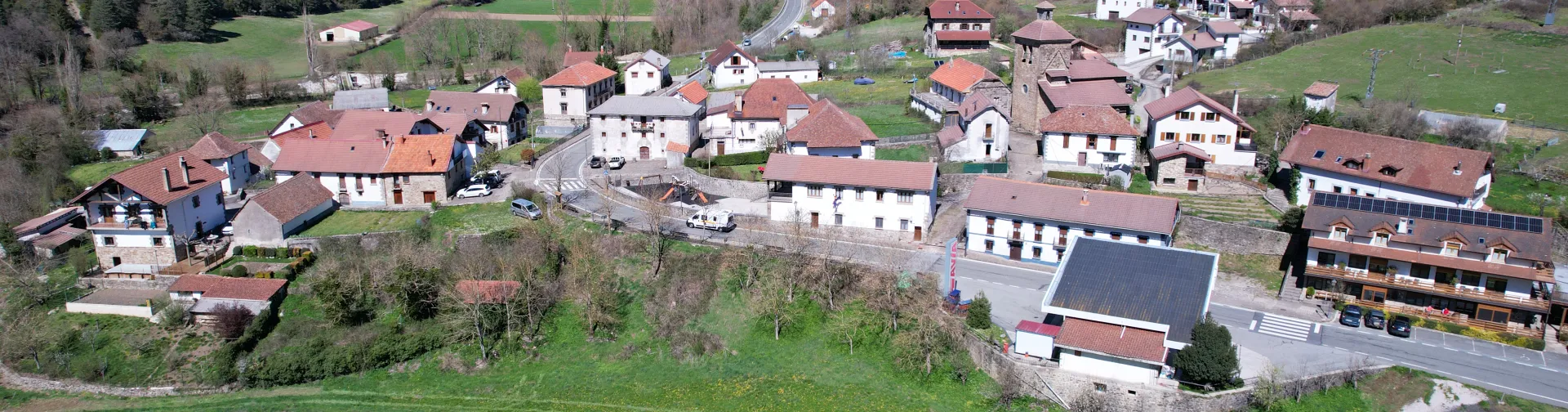 Vista aérea del pueblo de Oronz rodeado de montañas boscosas, casas con tejados rojos y prados verdes en el valle pirenaico.
