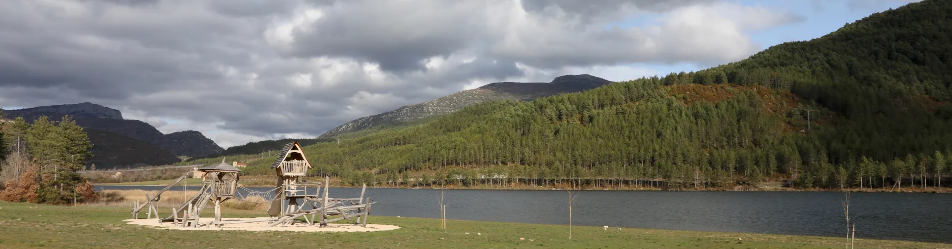 Embalse de Nagore rodeado de bosque pirenaico, con prado en primer plano y parque de madera y montes al fondo bajo cielo nublado.