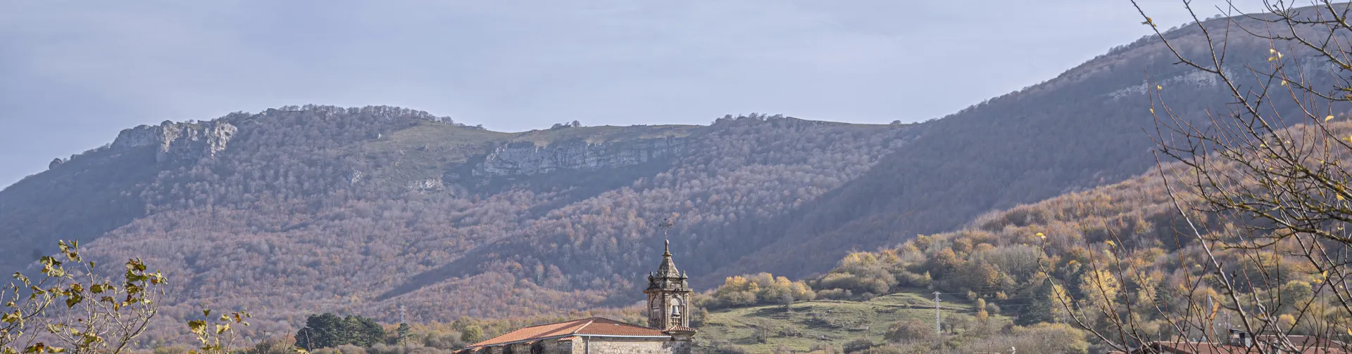 Iglesia con campanario sobre pueblo de Urdiain, rodeado de montañas boscosas y prados verdes.