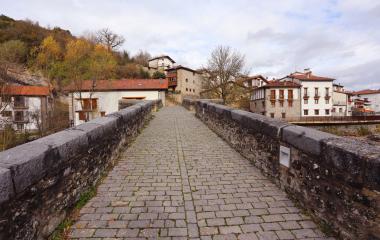 Puente de piedra empedrado sobre río en Burgui, con casas tradicionales de piedra y tejado rojo al fondo.
