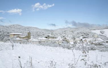 Pueblo de Erro cubierto de nieve con iglesia y montañas nevadas al fondo bajo cielo despejado.