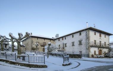 Plaza nevada de Erro con casas de piedra, fuente y árboles cubiertos de nieve bajo cielo despejado.