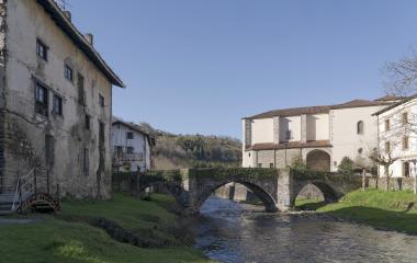 Puente de piedra medieval sobre río en Goizueta con la iglesia al fondo y pasarela de madera en primer plano.