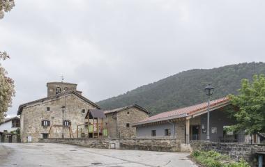 Iglesia  de piedra con campanario en plaza de Ilurdoz, anexa a parque infantil, con montaña de fondo.