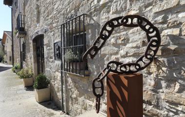 Escultura de cadena en hierro junto a fachada de piedra en Lizoain-Arriasgoiti, con ventana enrejada y calle empedrada.