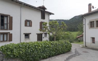 Calle con casas blancas con ventanas de madera en Loizu, Pirineo, rodeadas de vegetación y monte boscoso de fondo.