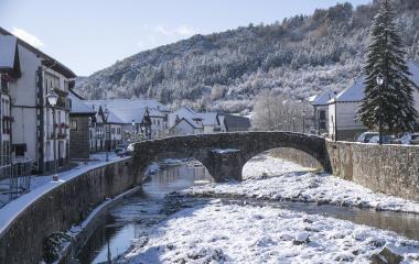 Puente de piedra sobre rio nevado en Ochagavia, con casas blancas y bosque al fondo.