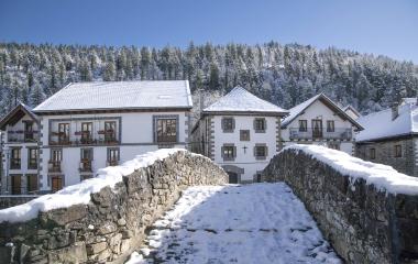 Puente nevado y casas tradicionales blancas de Ochagavía con bosque al fondo.