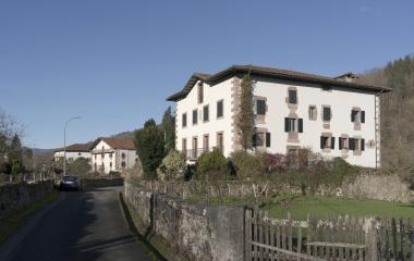 Casa blanca con balcón y portada, cercada por muro de piedra.