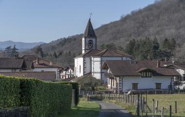 Vista general de Oronoz con una iglesia con campanario, casas al frente en ladera, prados y montes boscosos.