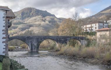 Vista de Sumbilla desde puente medieval de piedra con arcos sobre río, casas y montañas en la composición.