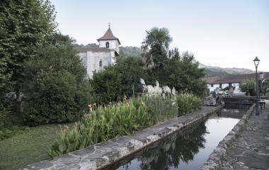 Canal de agua y  vegetación tropical en primer plano, con la iglesia entre los arboles de atrás en Urdax.