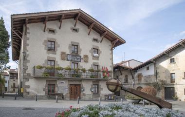 Ayuntamiento de piedra en Urdiain con balconada, escudos de piedra y bandera, delante una escultura de hierro.