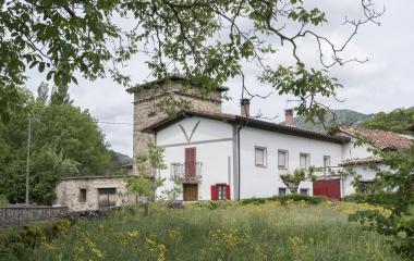 Casa con fachada blanca y edificio con torre de piedra en Uriz, Pirineo navarro, rodeada de vegetación verde y prado con flores amarillas.