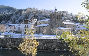 Iglesia de piedra con campanario en Urzainqui, rodeada de nieve con bosque pirenaico y río en primer plano.