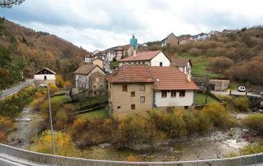 Pueblo de Uztarroz con casas de piedra junto a río, rodeado de bosque de montaña en otoño.