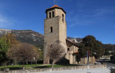 Iglesia de piedra con torre campanario de tejado rojizo en Yesa, rodeada de montes y vegetación al fondo.