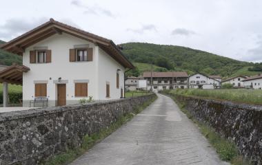 Conjunto de casas tradicionales en Zilbeti, con muros de piedra en camino que va al pueblo y monte boscoso de fondo.